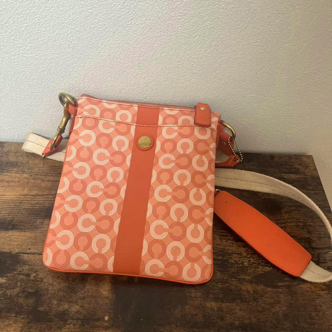 Orange Coach shoulder bag with repeating 'C' pattern, a vertical leather stripe and gold emblem, shown with a cream canvas shoulder strap on a wooden surface.