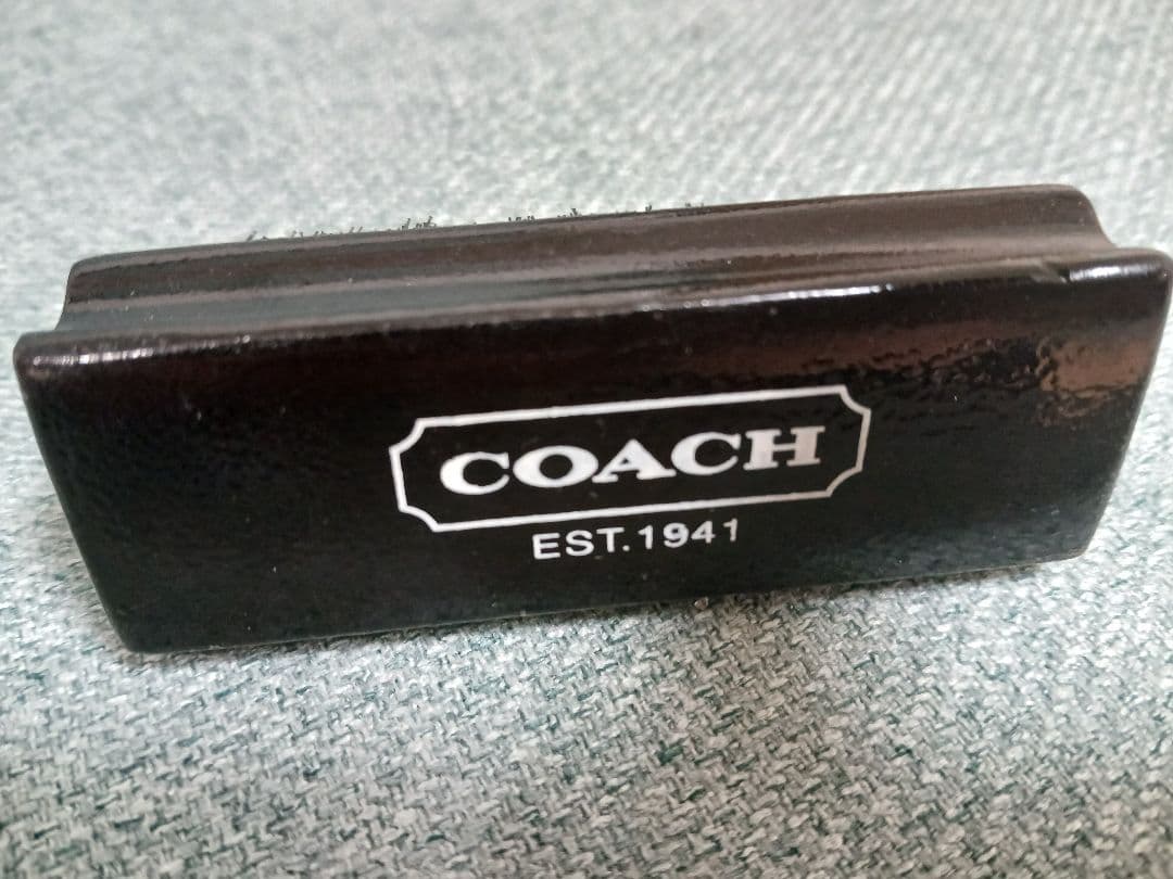 Black COACH cleaning brush showing the handle with the COACH logo and "EST.1941" on a fabric background.