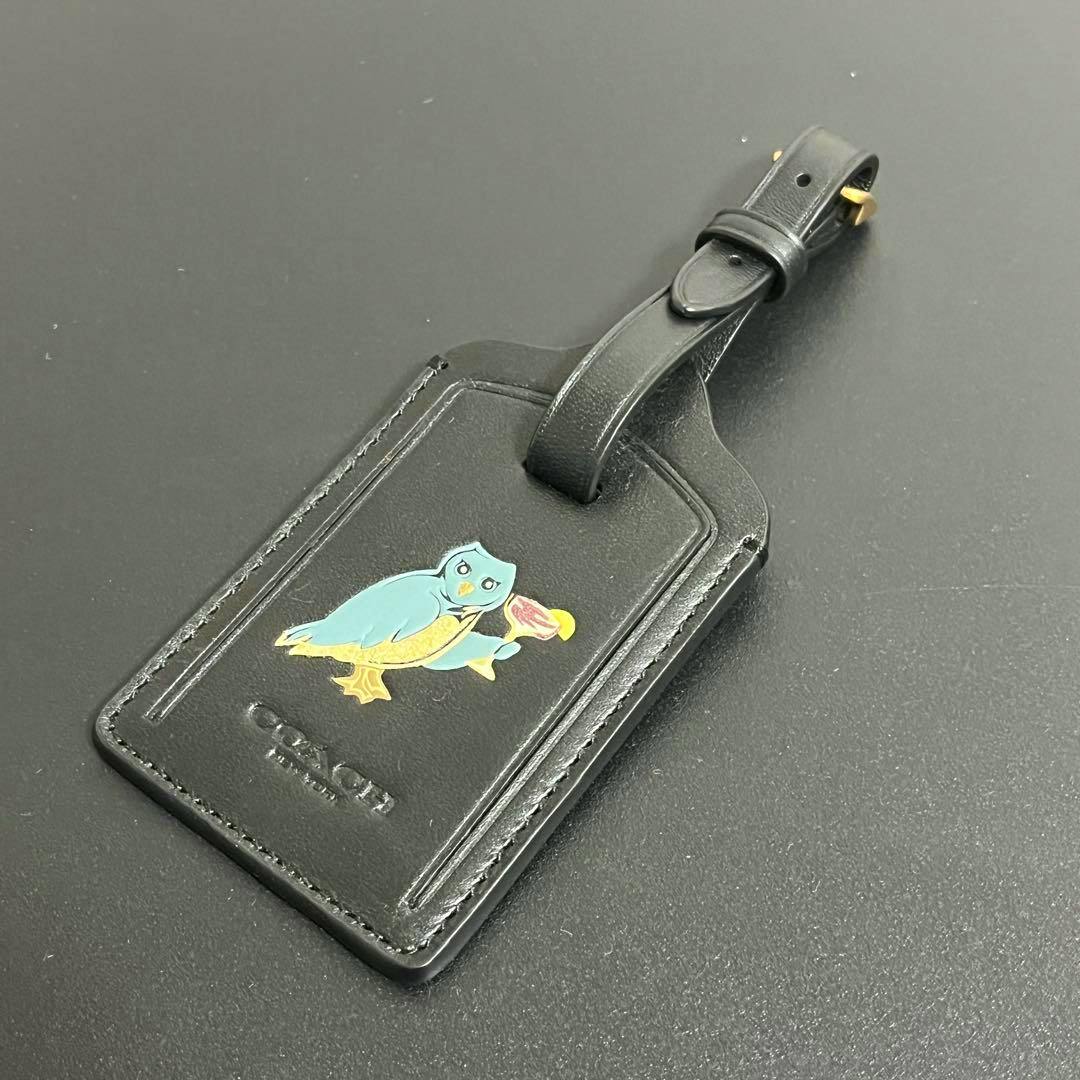 Black leather COACH luggage tag with a blue-and-yellow owl graphic and an attached buckle strap.