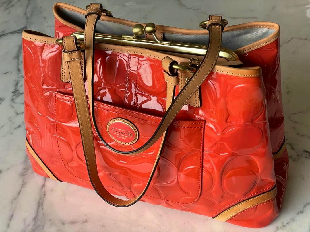Orange patent-leather COACH shoulder bag with tan handles and a front pocket, photographed on a marble surface.