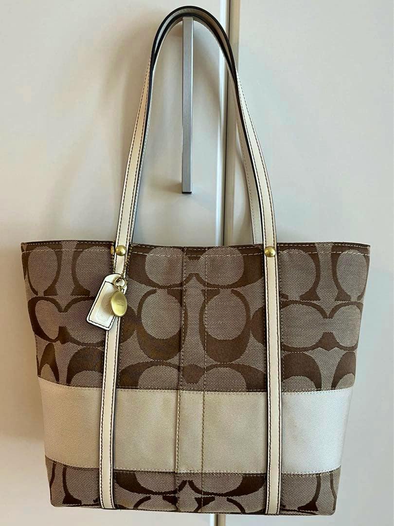 Front view of a Coach white and brown canvas handbag with signature C pattern, a white horizontal band, and white straps hanging on a hook.