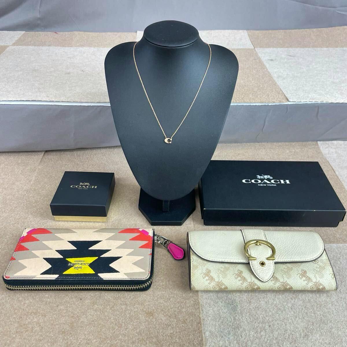 A long Coach wallet (right) and a patterned Coach zip wallet (left) displayed with a necklace on a black bust and Coach boxes behind.