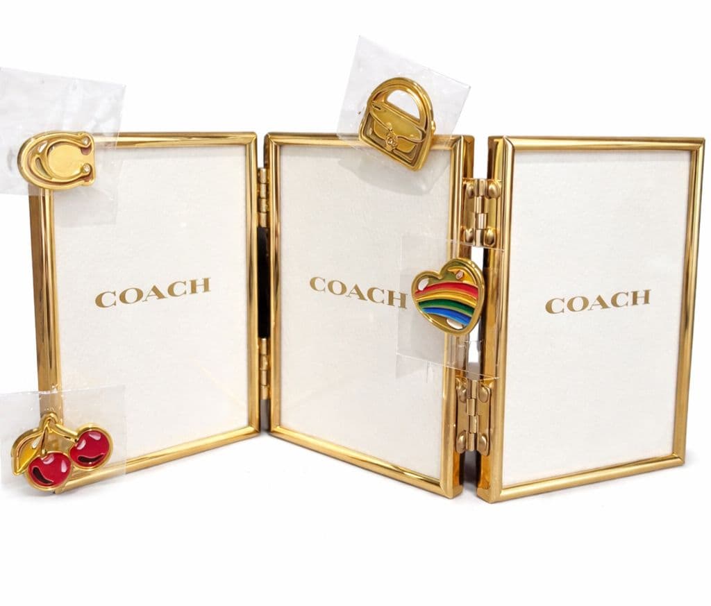 Front view of a gold-tone COACH triple-hinged photo frame with paper inserts and taped decorative charms.