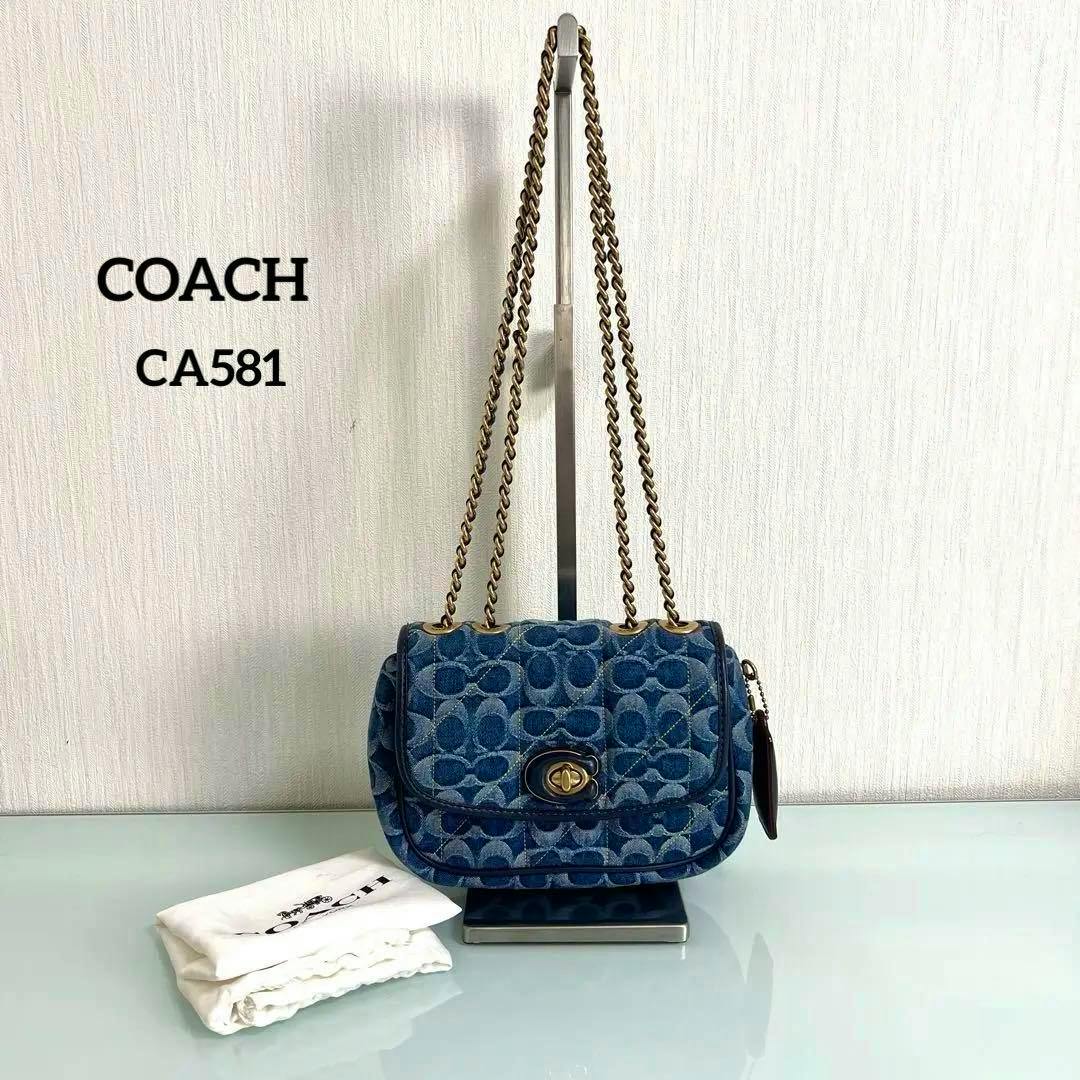 Denim Coach Pillow Madison CA581 shoulder bag with gold-tone chain strap shown on a display stand next to a folded dust bag.
