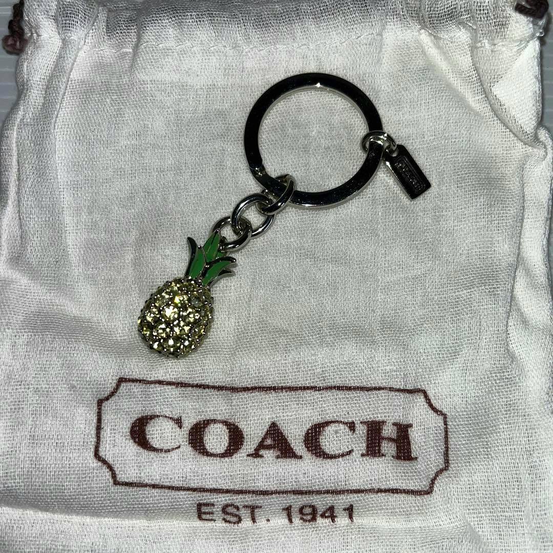 COACH pineapple bag charm keyring with green enamel leaves and yellow stones displayed on a white Coach dust bag.