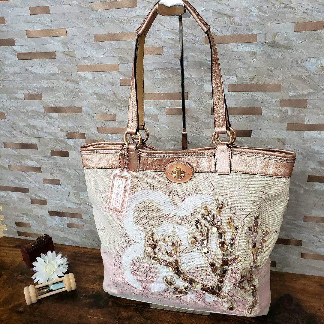 Front view of a COACH canvas pink tote bag with metallic handles and shell-style embellishment, displayed against a tiled background.