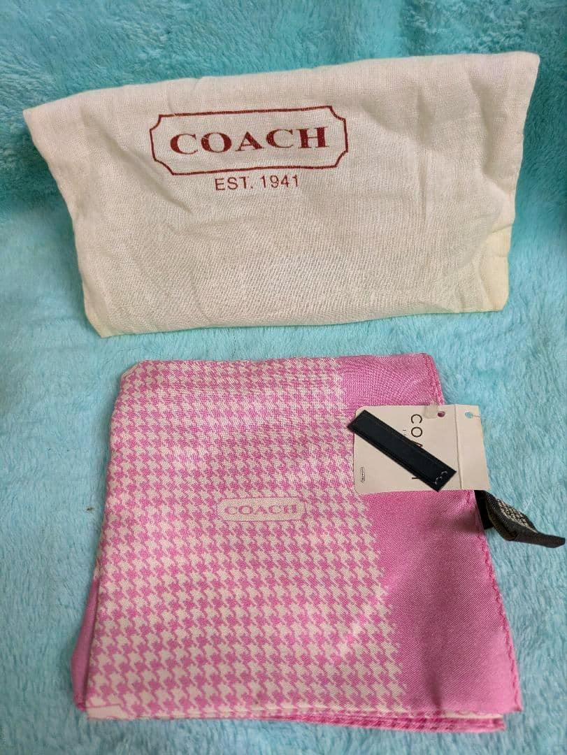 Folded pink houndstooth-pattern COACH scarf with tags on a blue surface and a COACH dust bag behind it.