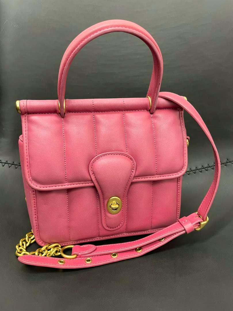 COACH pink leather shoulder bag with top handle, front flap with a gold turn-lock, and a chain-and-leather shoulder strap against a dark background.