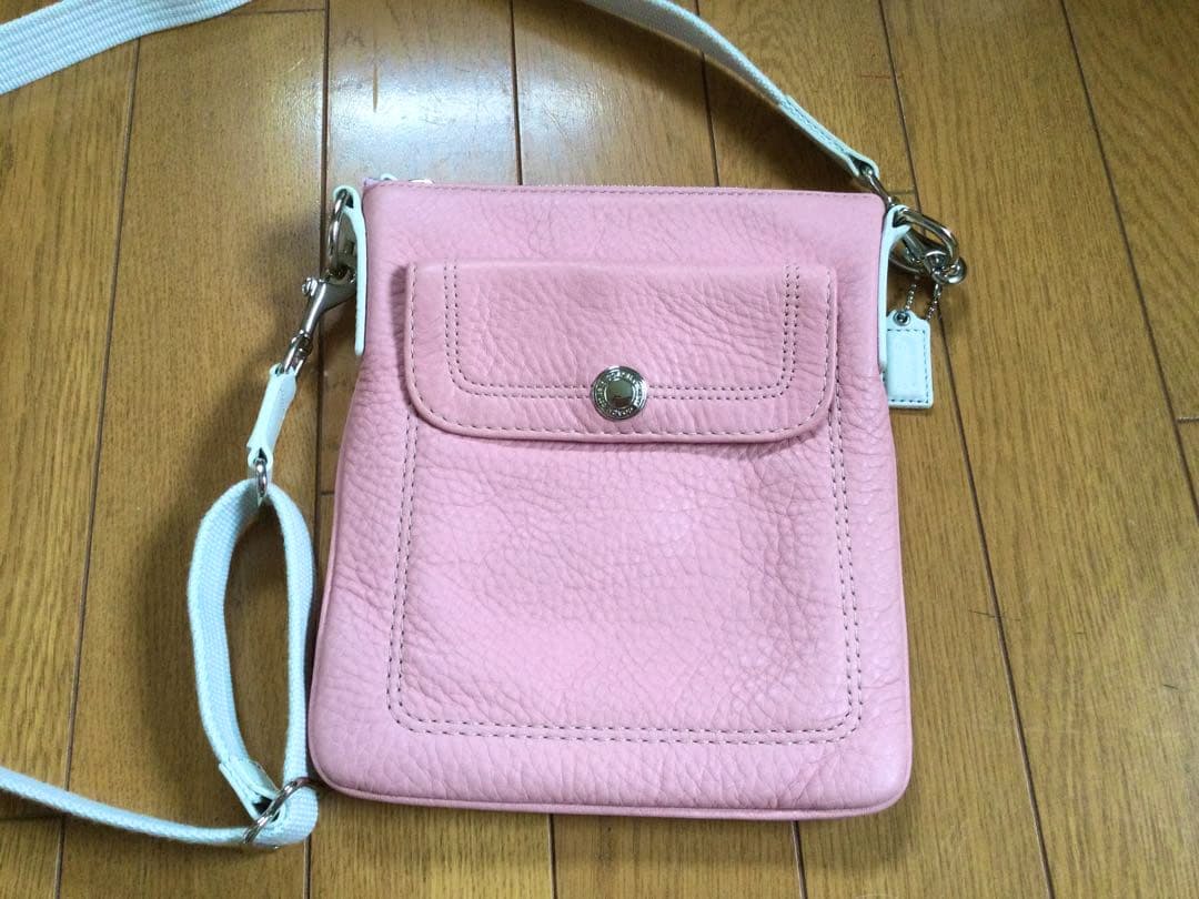 Front view of a pink Coach leather shoulder bag with a front flap pocket and white shoulder strap on a wooden floor.