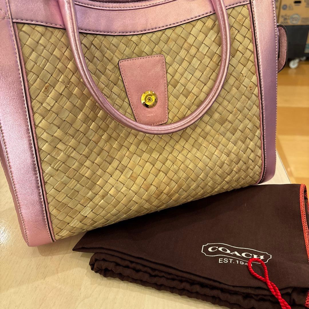 Front view of a straw basket COACH handbag with pink leather trim and handles shown with a COACH dust bag.