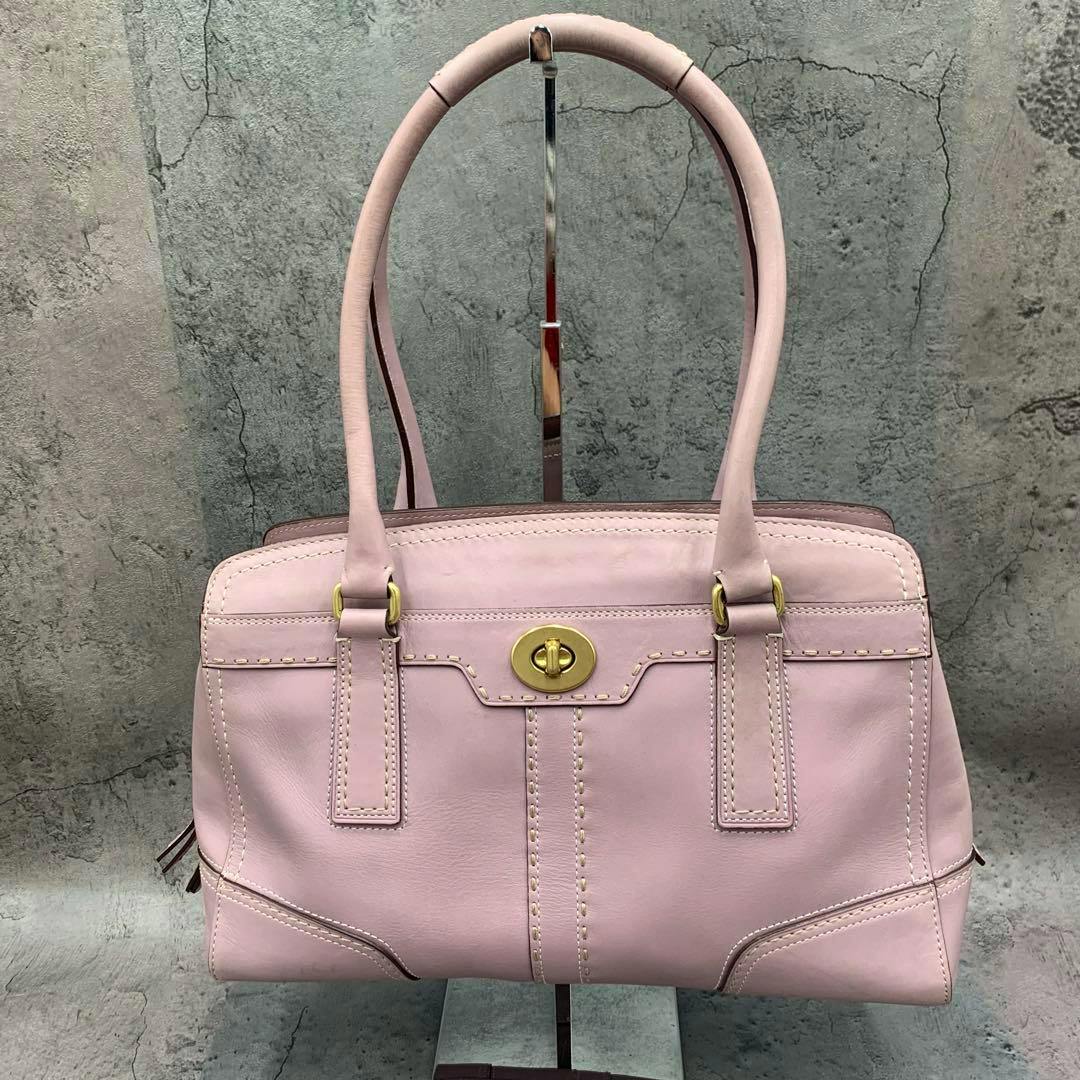 Front view of a pink COACH turn-lock leather handbag with double top handles displayed on a stand.