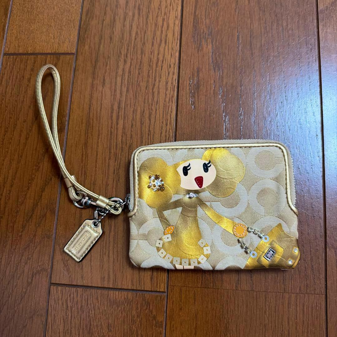 Gold Coach signature canvas coin case with a Poppy character print and matching wrist strap on a wooden floor.