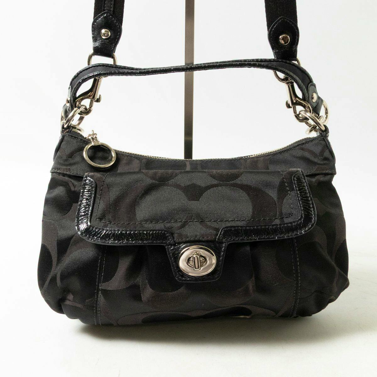 Front view of a black COACH signature-pattern 2-way shoulder bag with a front flap pocket, turn-lock closure, and short handles.