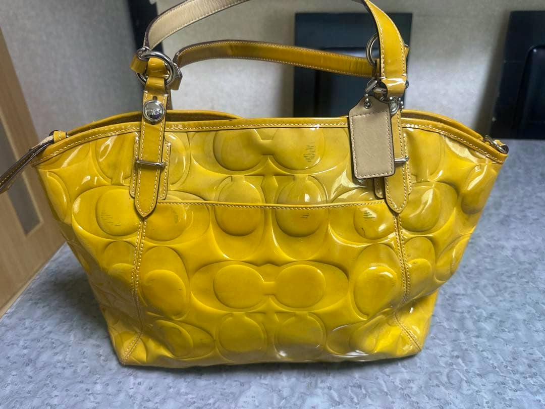 Front view of a yellow COACH Poppy tote bag (F13759) in patent leather with embossed pattern and top handles on a gray surface.