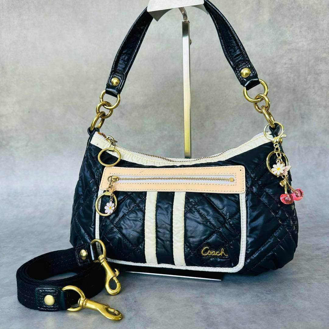 Black quilted Coach Poppy cow leather 2-way bag with cream trim, gold hardware, floral and cherry charms, and a detachable strap on a display stand.