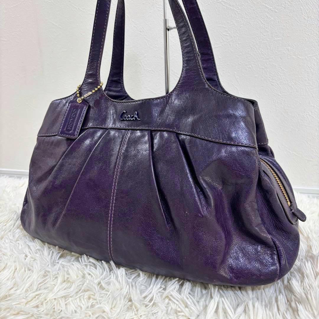 Front view of a purple COACH leather tote (hobo-style) bag with double shoulder straps and a hanging Coach tag, placed on a white rug.