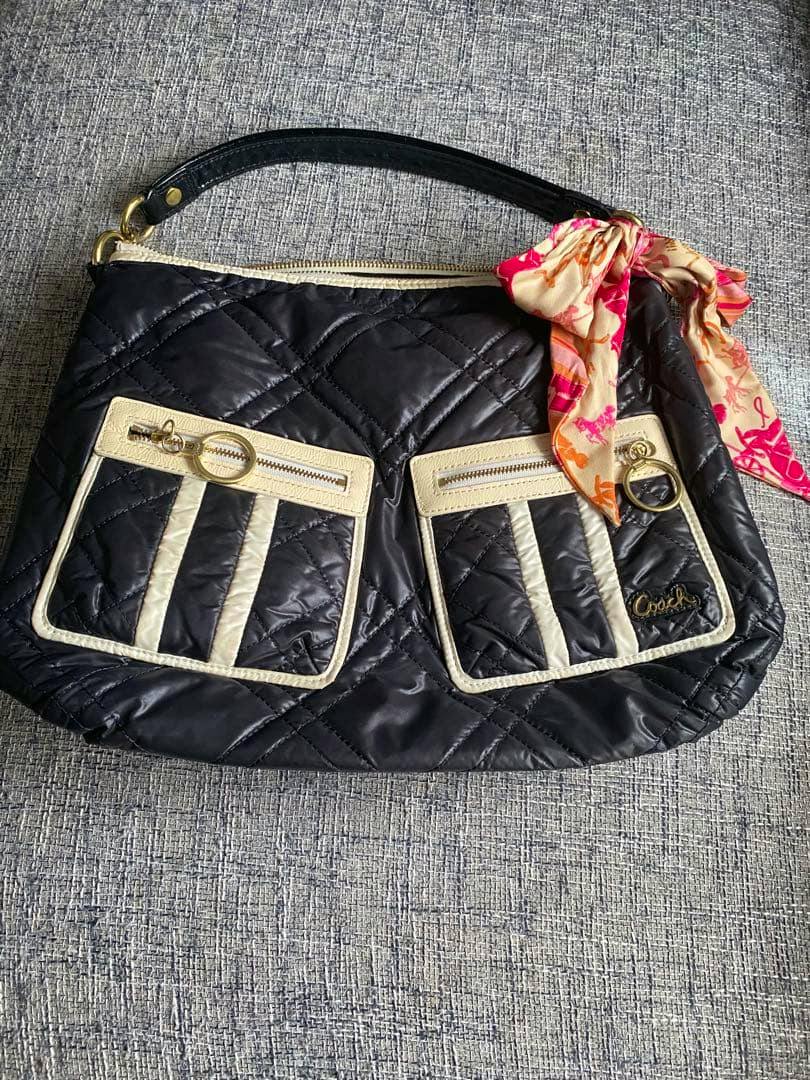 Front view of the COACH black quilted shoulder bag (no shoulder strap) showing two front pockets with cream trim, a short handle, and a pink patterned scarf tied to the hardware.