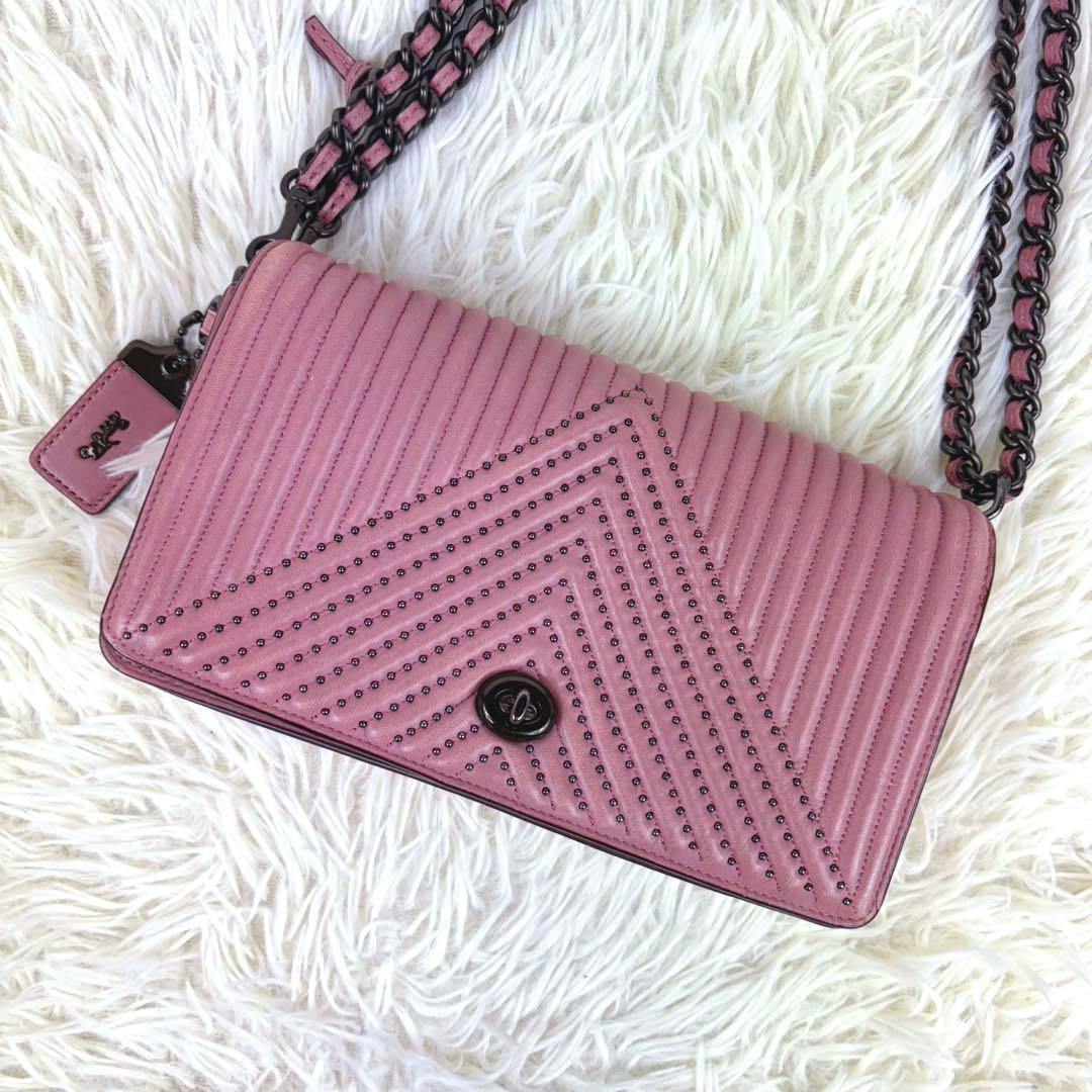 Pink quilted leather Coach shoulder bag with chevron stud detailing, chain strap, and turn‑lock closure on a white faux‑fur background.