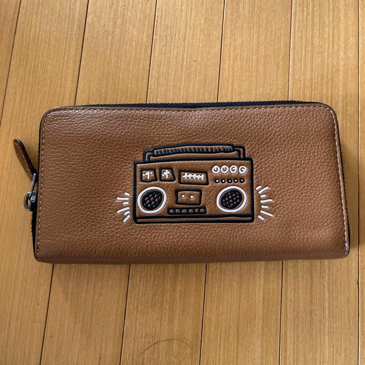 COACH brown long zip-around leather wallet with a radio/cassette motif on the front, pictured on a wooden floor.