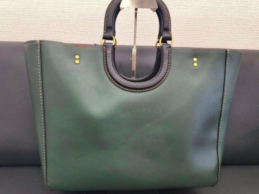 Front view of a dark green Coach leather tote bag with circular top handles and gold-tone hardware.
