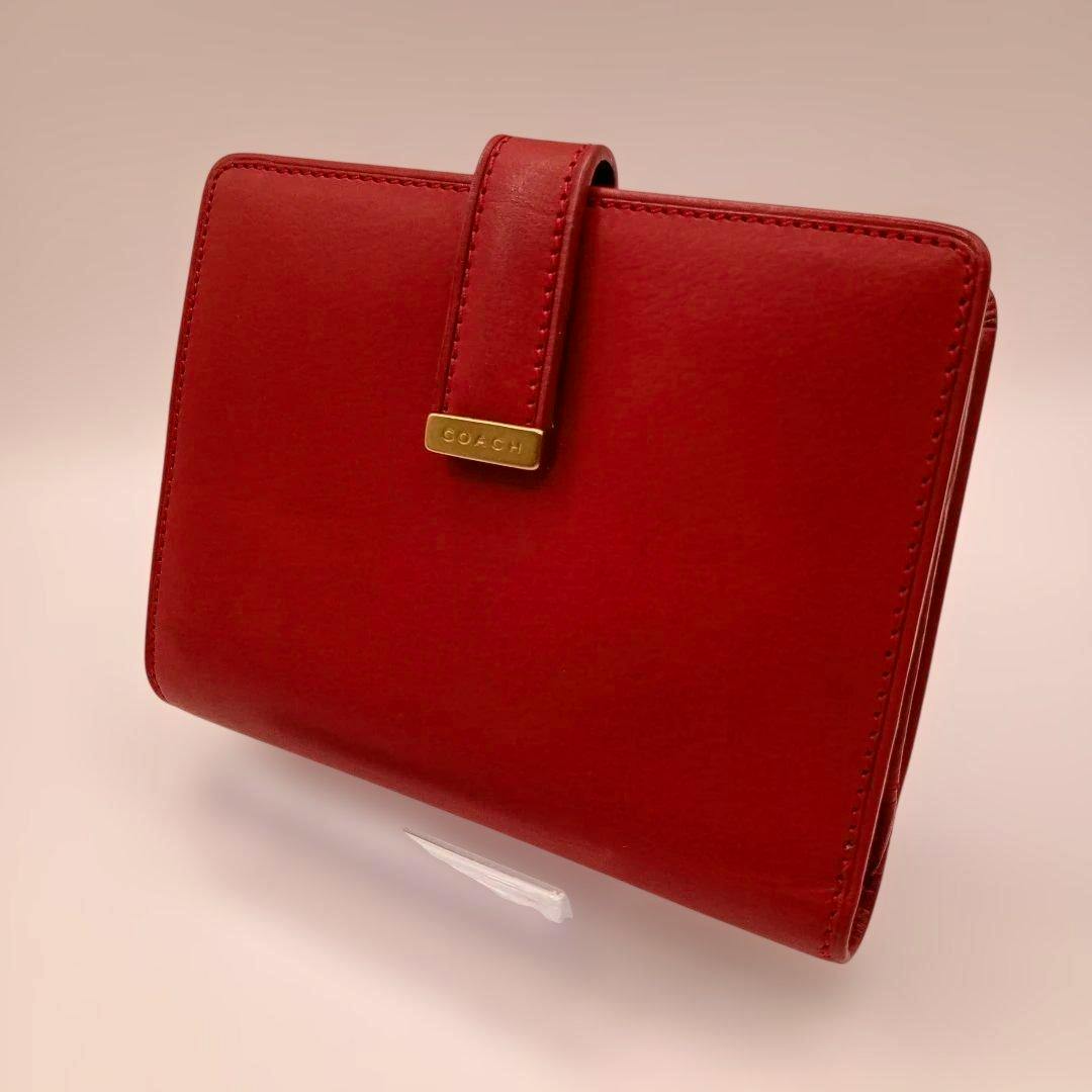 Red COACH bi-fold leather wallet shown closed with a stitched strap and gold COACH plaque visible on the front.