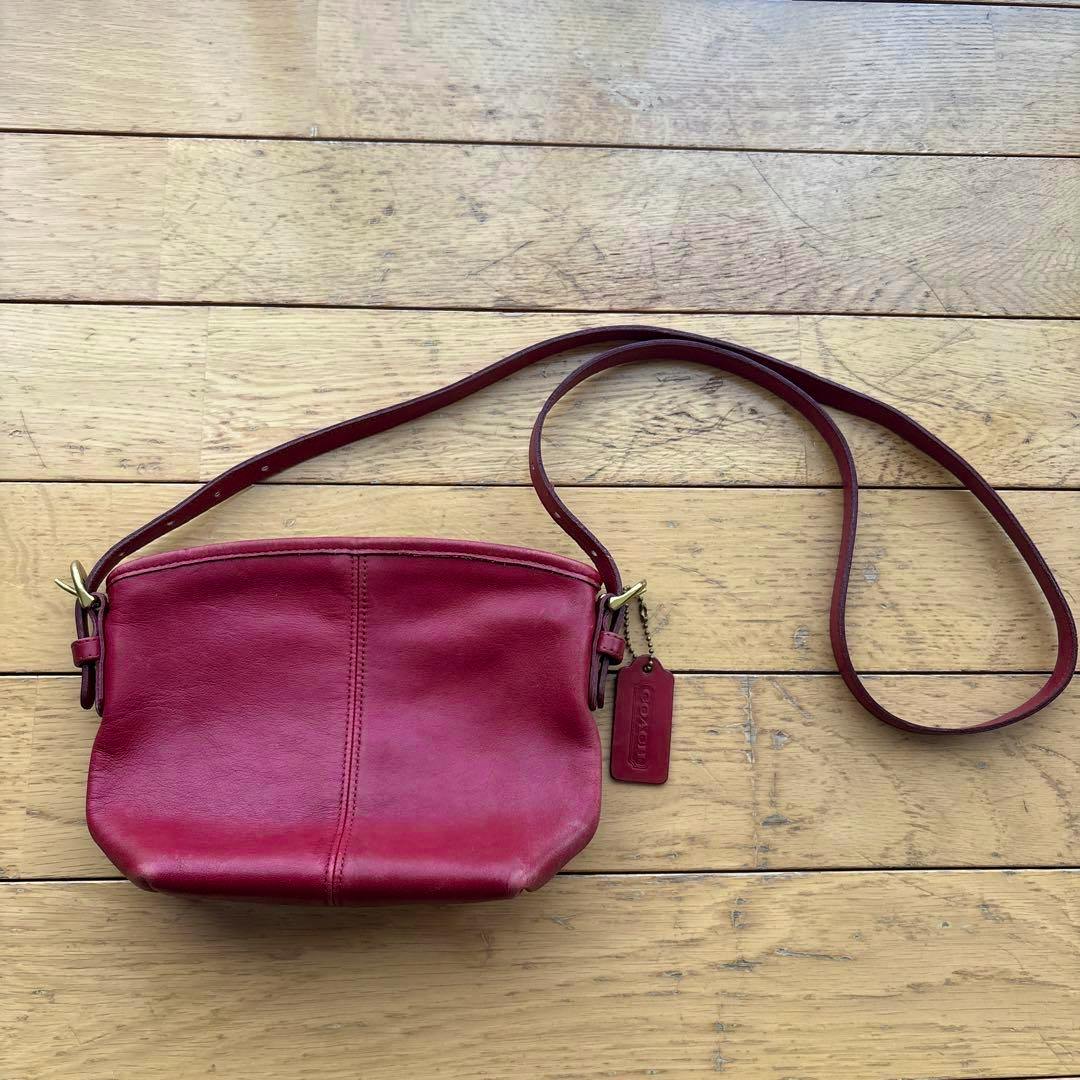 Red Coach leather shoulder bag with long strap and leather tag lying on a wooden floor.