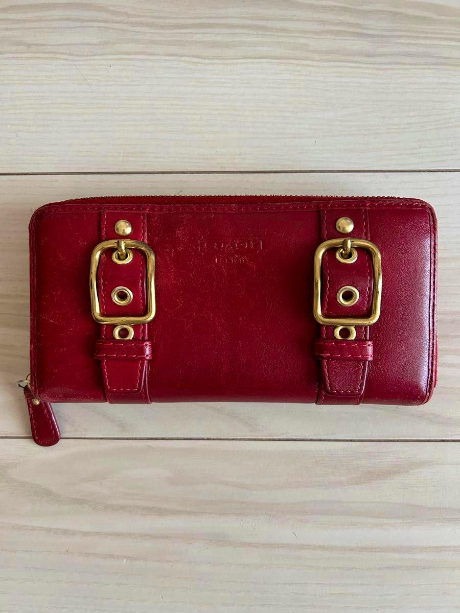 Red COACH long leather zip-around wallet with belt-style buckle accents on the front, photographed on a light wood surface.