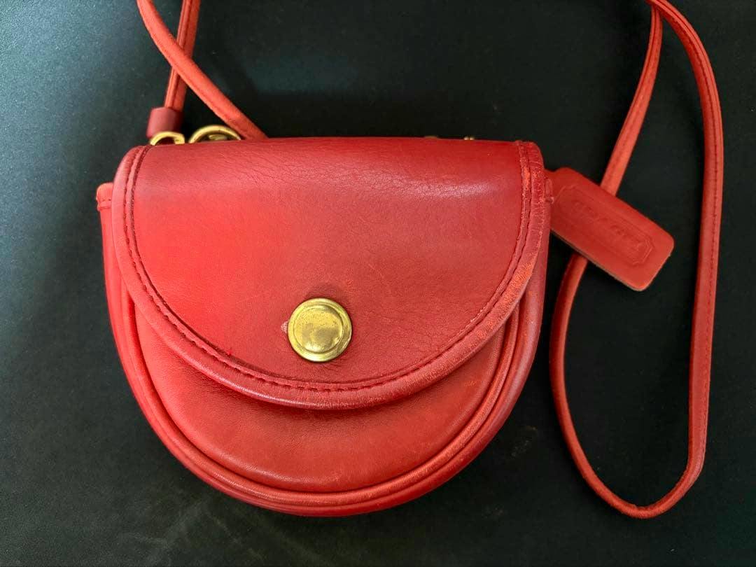 Red Coach leather mini shoulder bag with a flap and round gold-tone button, visible strap and leather tag on a black background.