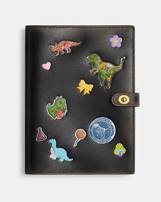 Front of a cow leather notebook cover with colorful dinosaur sticker print and a snap strap closure.