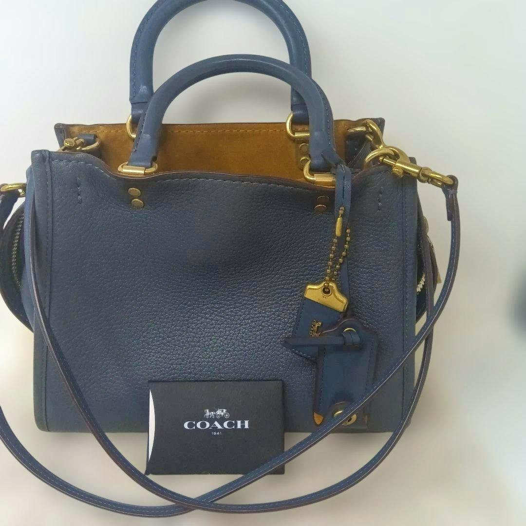 Blue leather COACH 2-way bag with top handles, a shoulder strap, leather hangtag, and a small COACH card placed in front.