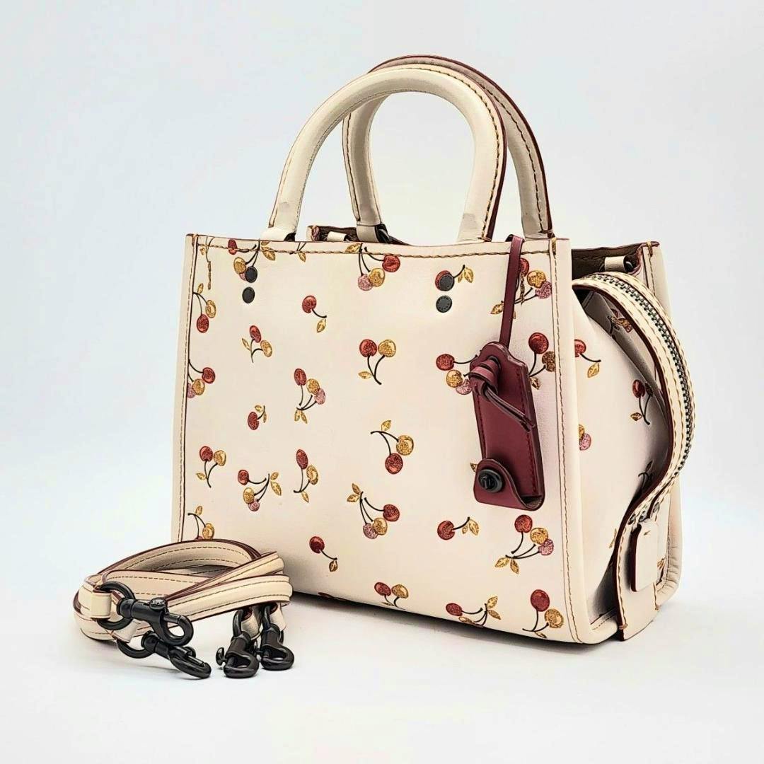 Cream-white Coach Rogue25 cow leather bag with cherry print, top handles, a detachable strap beside it, and a red leather tag visible.