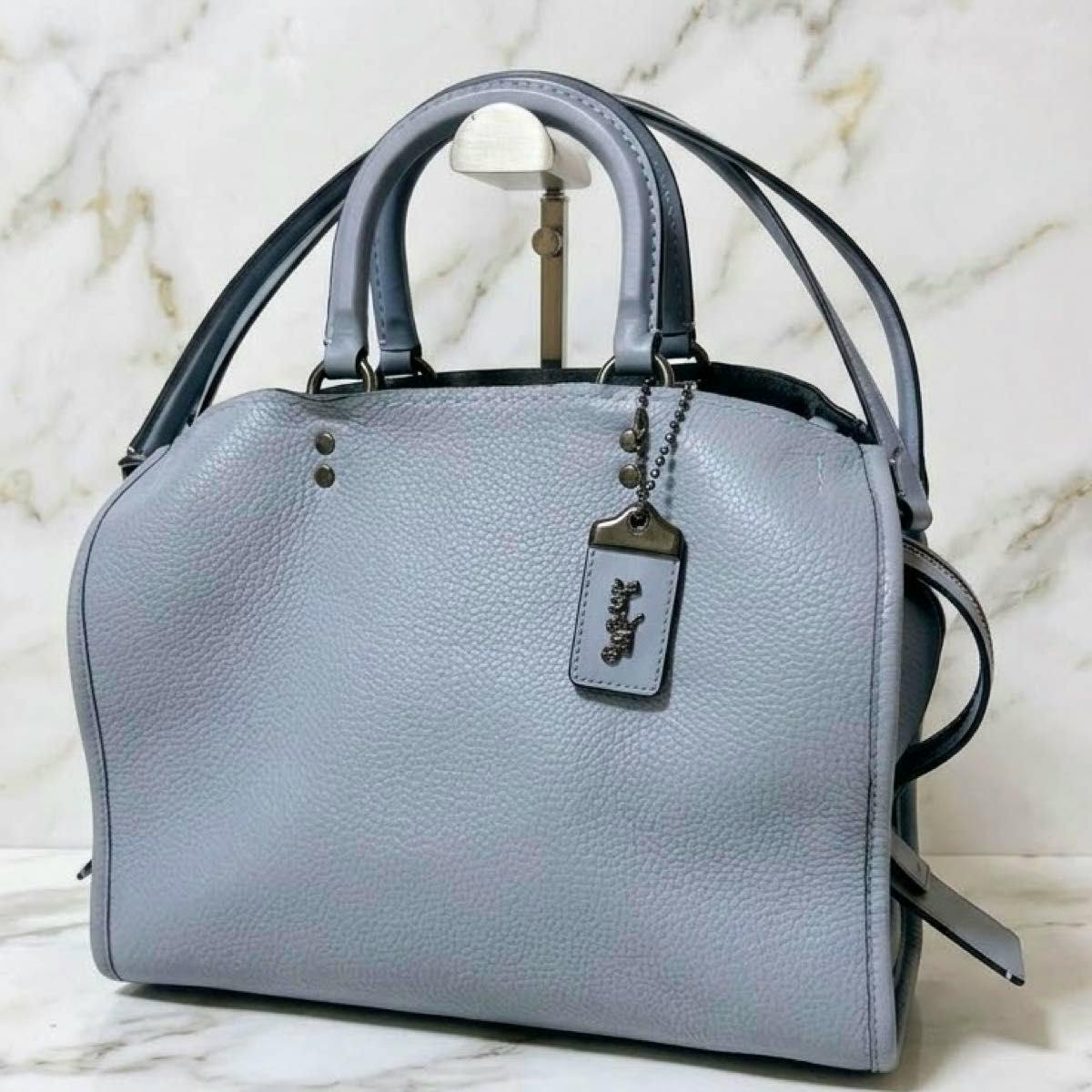 Light gray pebbled leather COACH Rogue handbag with top handles, shoulder strap, and logo hangtag on a display stand.