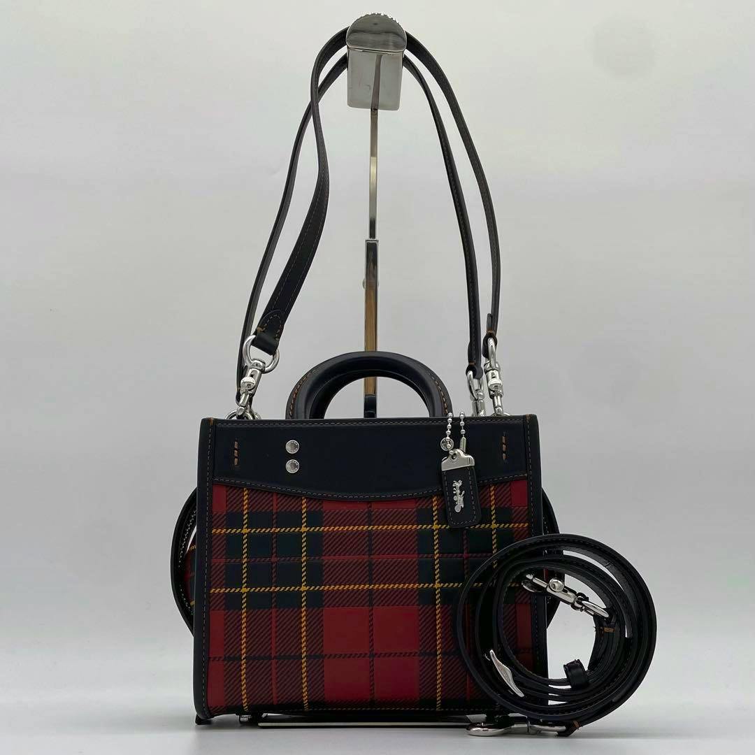 Red and black check-patterned Coach 3‑way shoulder bag with leather trim and detachable straps displayed on a stand.