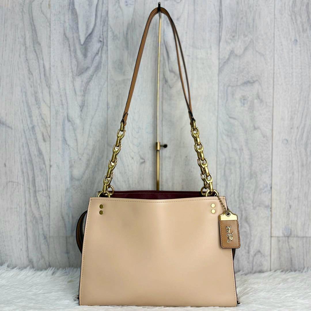Beige COACH leather tote bag with gold chain-and-leather shoulder straps and a leather hang tag displayed against a light wood background.