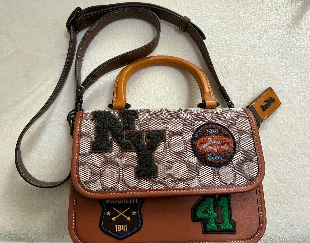 Front view of a Coach top-handle signature textile bag with brown leather trim, patch appliqués, and a detachable shoulder strap.