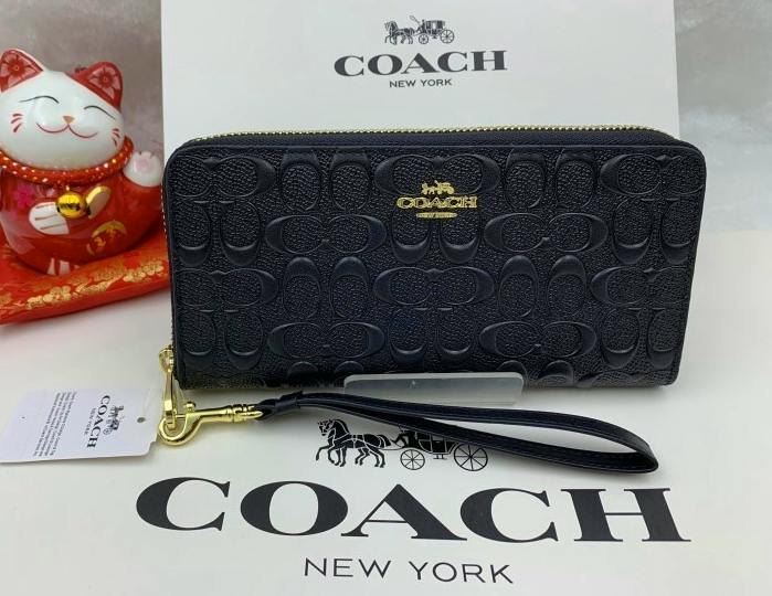 Black COACH long zip-around wallet with embossed signature pattern, gold-tone logo and wrist strap shown on a COACH-branded background.