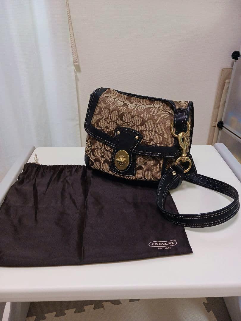 Beige and black Coach shoulder bag with monogram pattern displayed on a table alongside its black Coach dust bag.