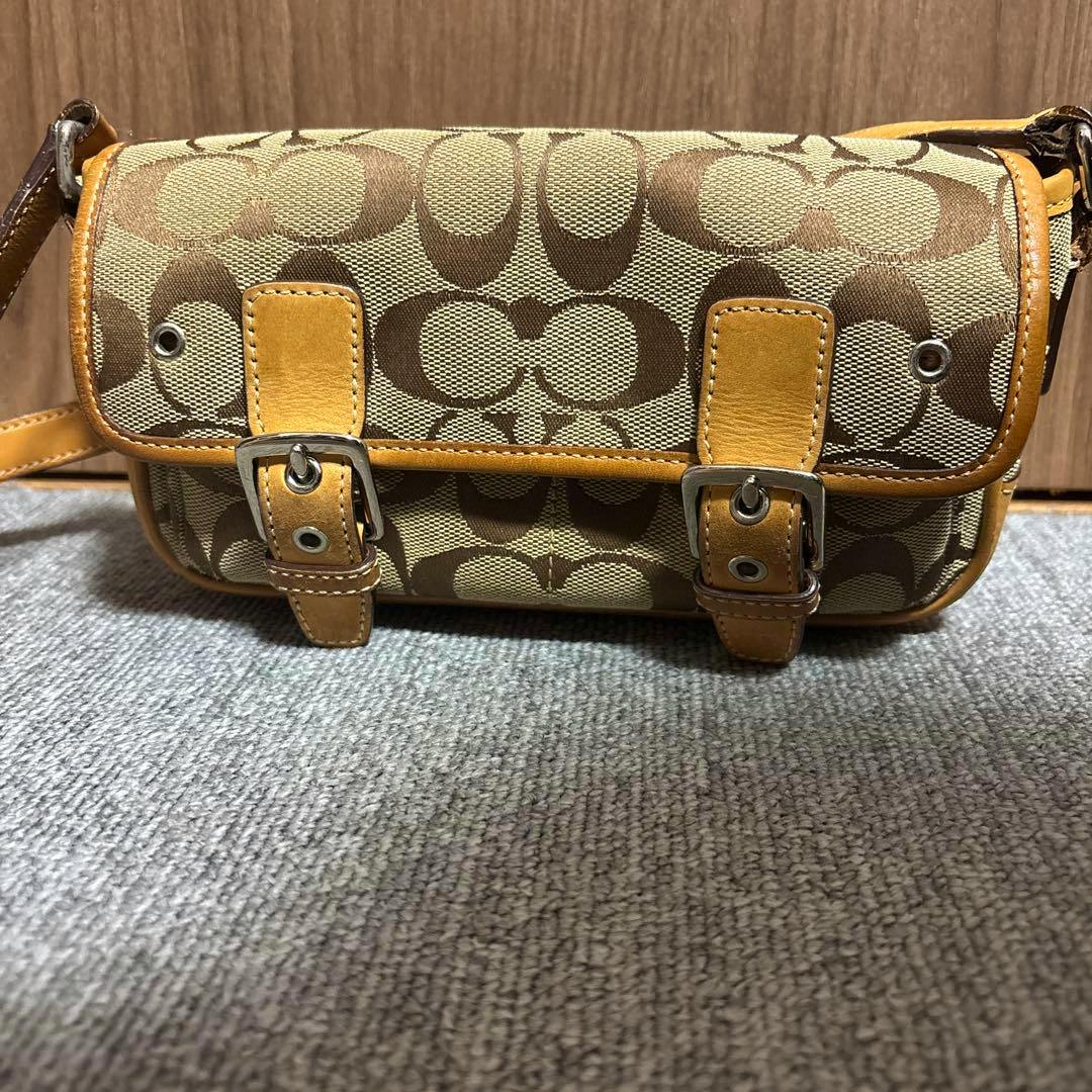 Front view of a brown and beige Coach canvas shoulder bag with signature 'C' pattern and two leather buckle straps.