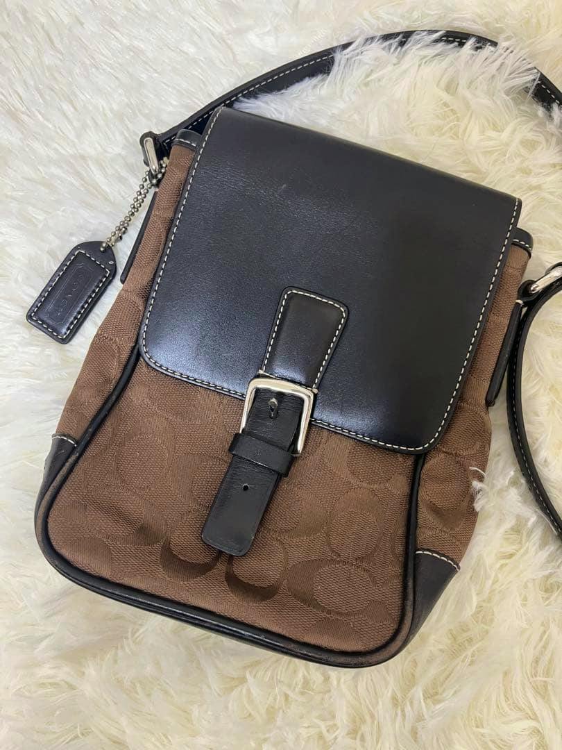 Brown Coach canvas shoulder bag with a black leather flap, buckle, and strap shown on a white fur background.