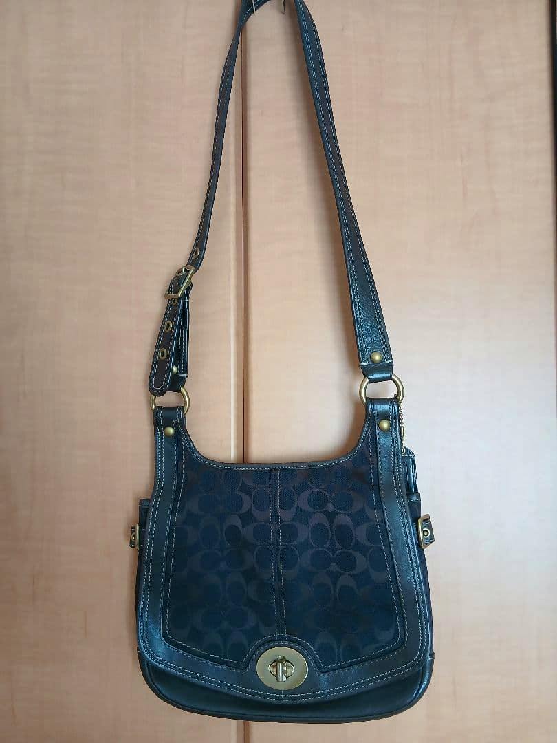 Front view of a dark Coach shoulder bag with signature C pattern, leather trim, strap, and a turn-lock closure hanging against a door.