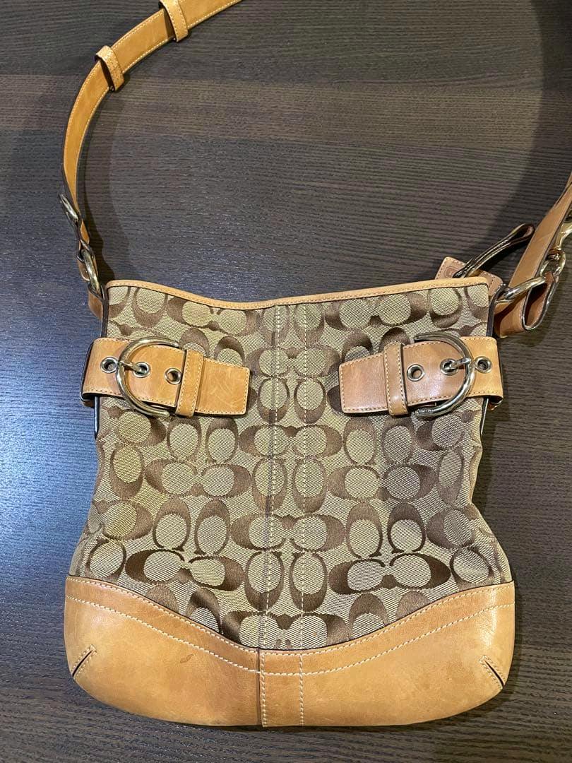 Front view of the Coach signature 2-way canvas shoulder bag with tan leather trim and buckle details on a dark surface.