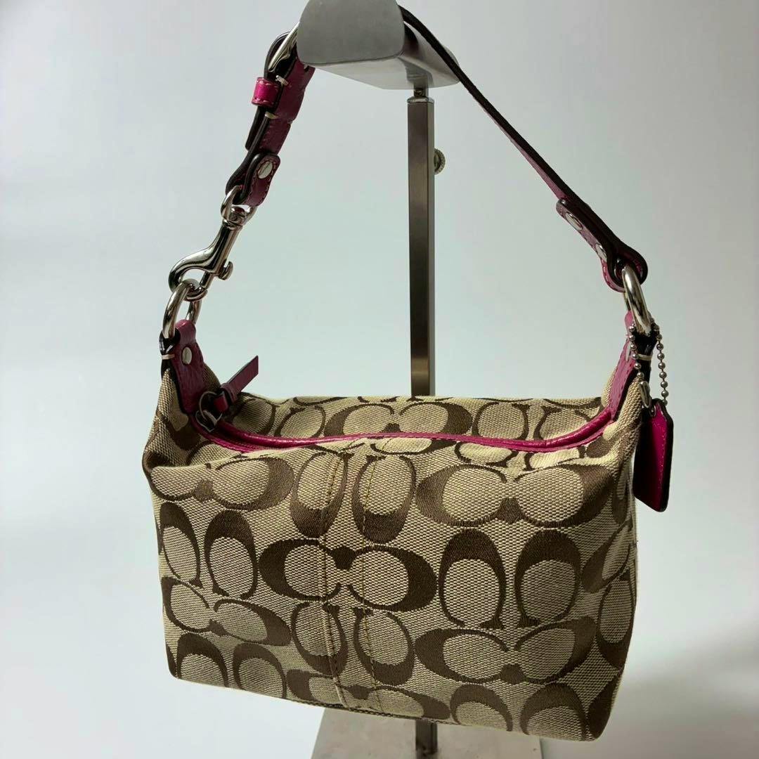 Beige and brown Coach signature fabric accessory pouch with pink leather trim and short strap hanging on a display stand.