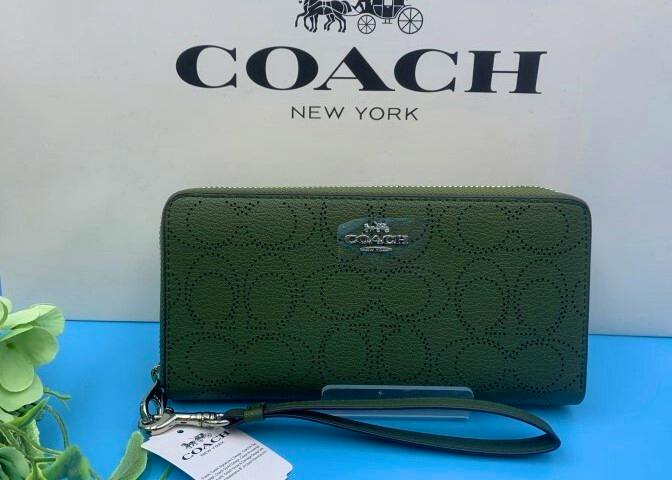 Green Coach long zip-around wallet with wrist strap and metal logo displayed against a Coach backdrop.