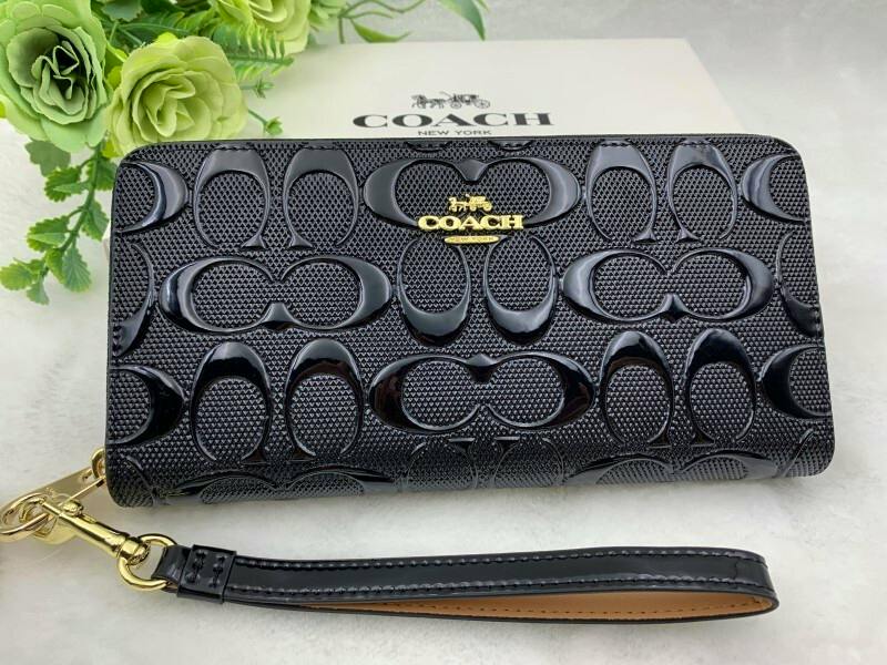Black COACH long zip wallet with signature embossed pattern and a detachable wrist strap on a light surface.