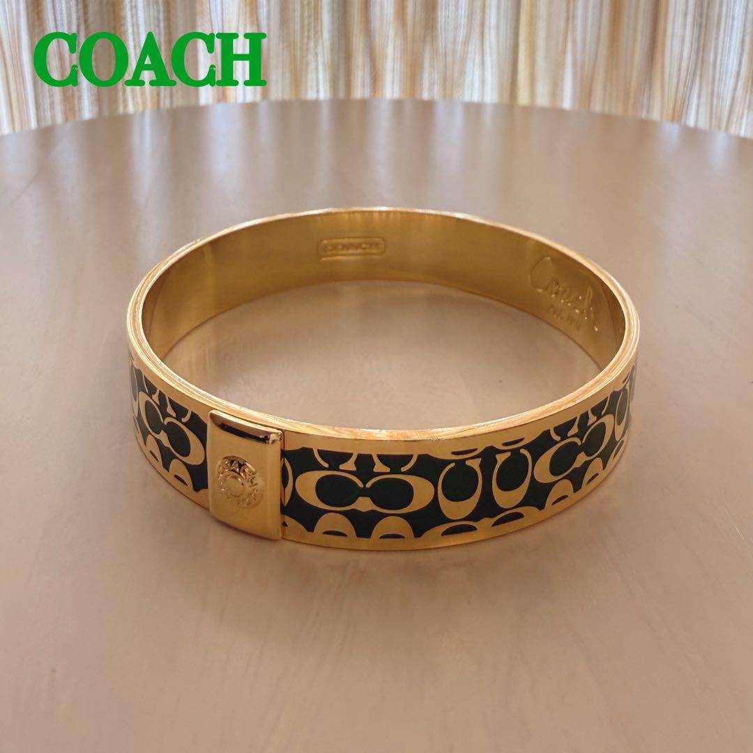 Gold bangle bracelet with Coach signature green pattern and a gold clasp resting on a tabletop.