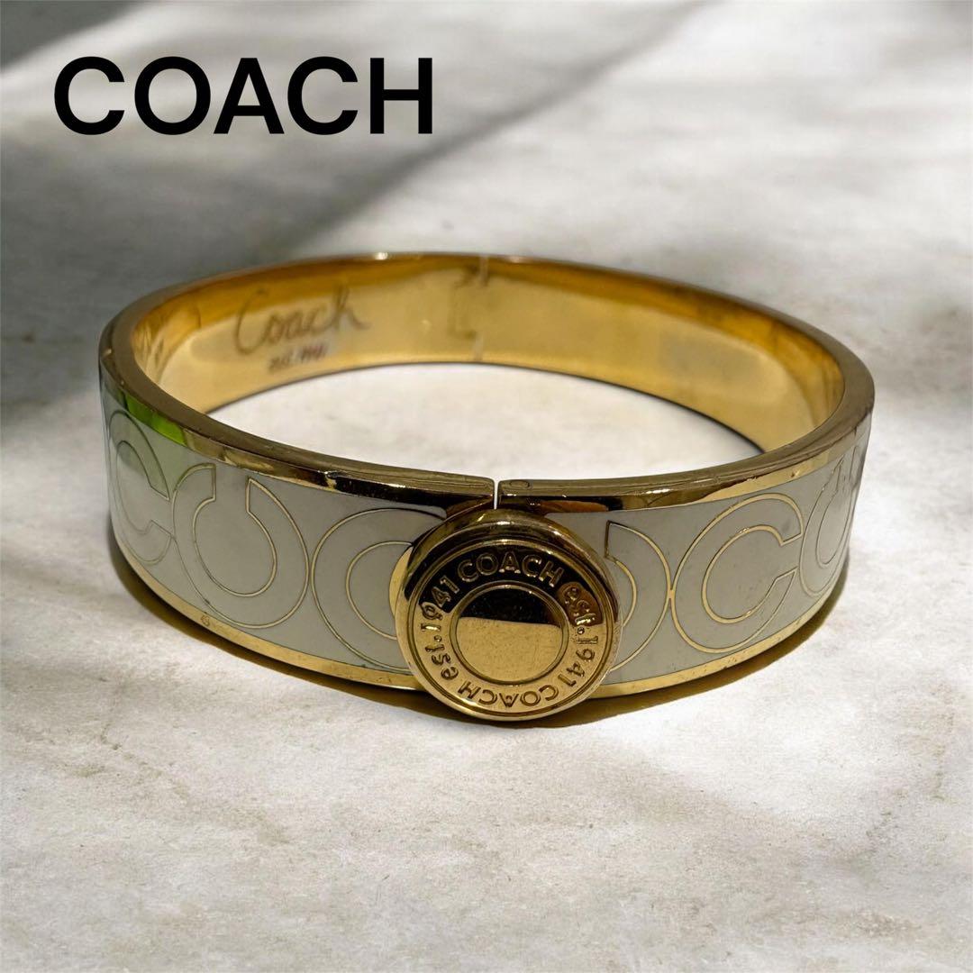 Gold-tone Coach signature bangle bracelet with white enamel detailing and circular logo clasp photographed on a light surface.