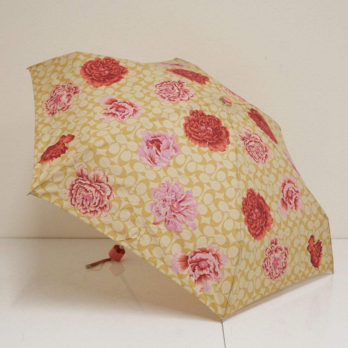 Beige Coach folding umbrella with signature 'C' pattern and pink and red floral print, shown open on a white background.