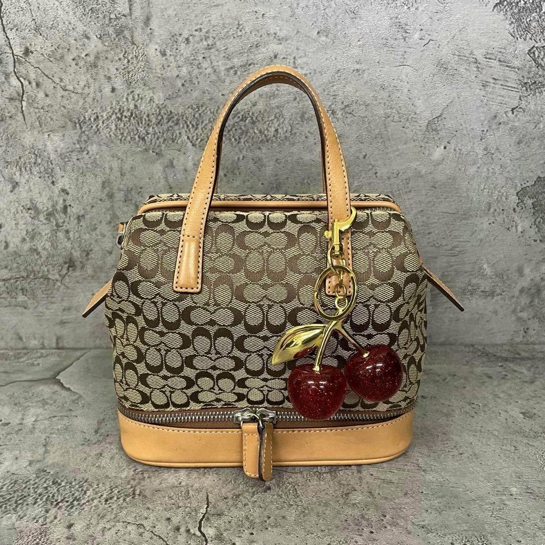 Front view of a beige Coach signature canvas handbag with leather trim, zipper closure, and a decorative cherry keychain.