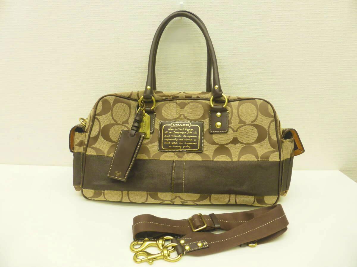 Front view of a beige Coach signature canvas Boston travel bag with top handles and a brown removable shoulder strap placed in front.