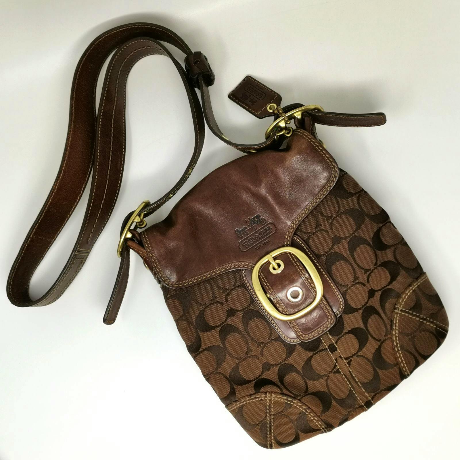 Brown Coach signature canvas shoulder bag with leather flap, gold buckle, and long strap on a white background.
